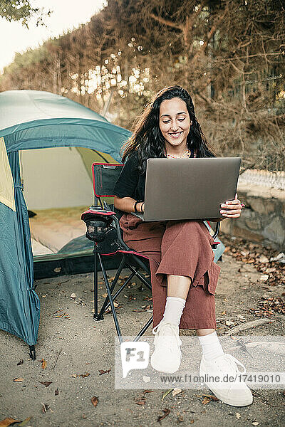 Smiling young woman using laptop while sitting on chair in forest