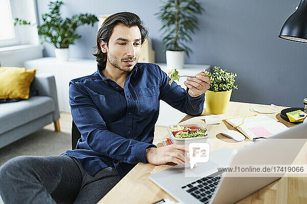 Businessman eating lunch while working on laptop at home