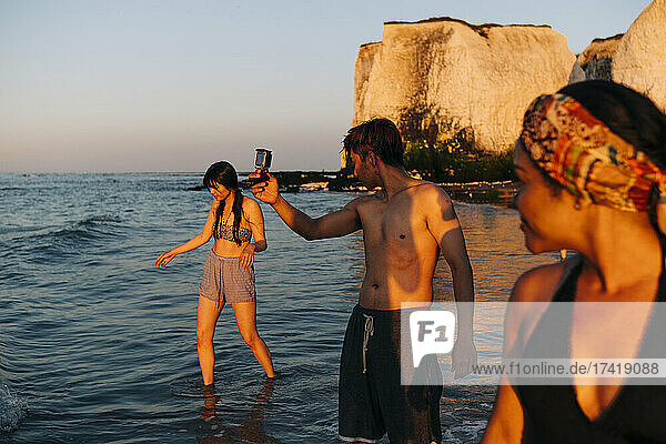 Male and female friends looking at woman walking through sea water during sunset