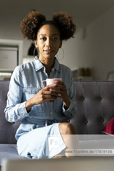 Afro young woman holding coffee cup sitting on sofa at home