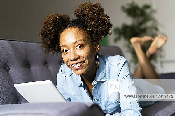 Smiling Afro woman with digital tablet lying on sofa in living room