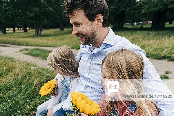 Cheerful father with arms around daughters sitting at park