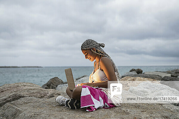 Smiling young woman using laptop on rock at beach during sunset