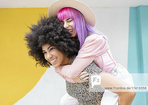 Afro woman piggybacking cheerful female friend in front of wall
