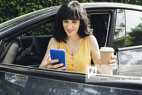 Glückliche Frau mit Einwegkaffeetasse und Smartphone beim Aussteigen aus dem Elektroauto