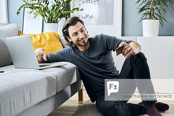 Smiling man with credit card sitting near sofa at home