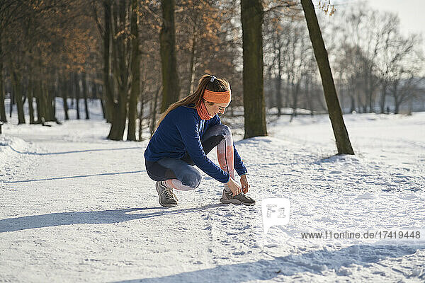 Young woman tying shoelace while crouching on snow during winter