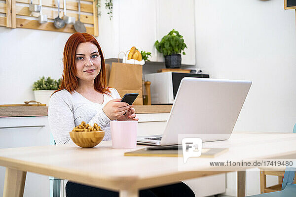 Smiling redhead woman with mobile phone and laptop sitting at home