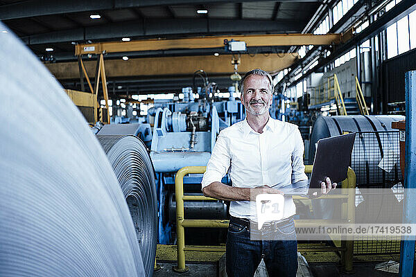 Smiling male business professional holding laptop while standing at steel mill