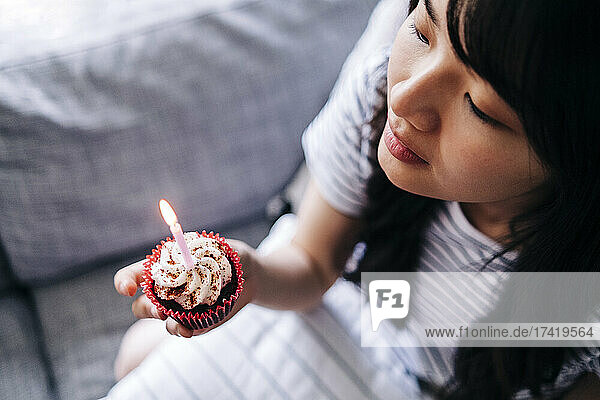 Junge Frau hält Cupcake und Kerze in der Hand  während sie auf dem Sofa sitzt