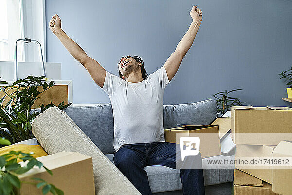Happy man with arms raised sitting on sofa amidst cardboard boxes at home