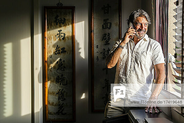 Smiling man looking through window while talking on mobile phone at home
