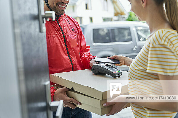Woman paying through mobile phone while receiving pizza order from deliveryman at doorway
