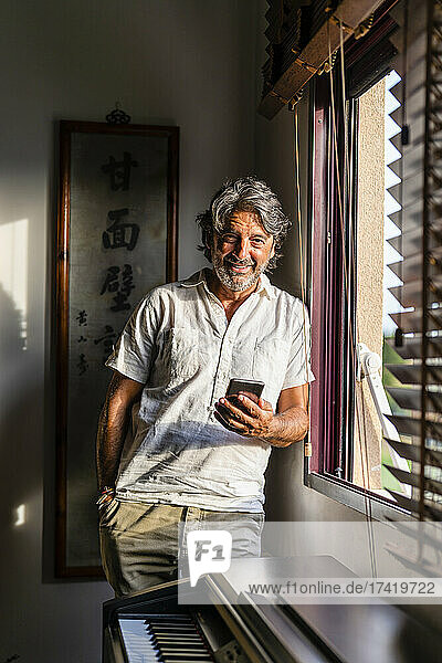 Smiling man with mobile phone standing by window
