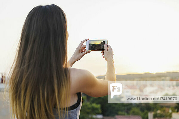 Woman photographing sunset view through smart phone at rooftop