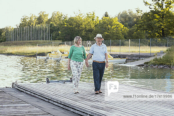 Älteres Paar hält sich an den Händen  während es auf dem Steg am See spaziert
