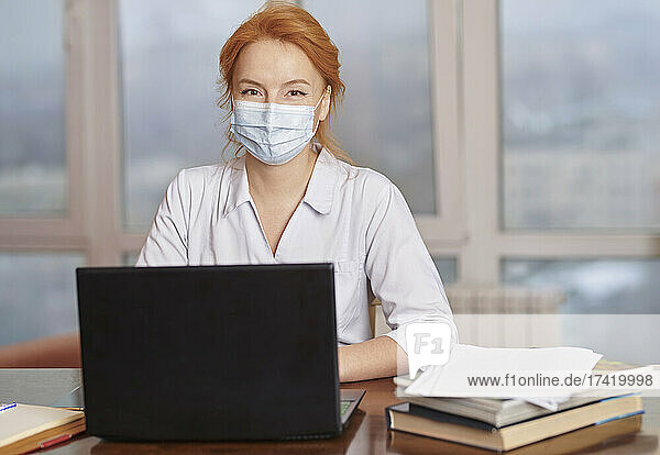 Female healthcare worker wearing protective face mask sitting with laptop in hospital