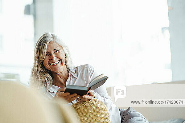 Happy mature woman sitting with book on chair in cafe