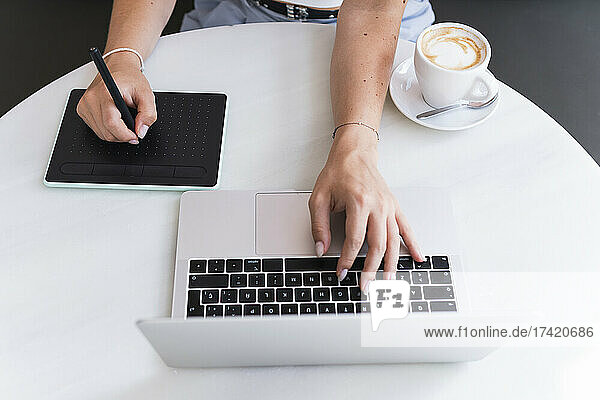 Female design professional sitting with laptop and graphic tablet in cafe