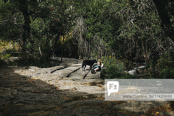 Dog standing by female owner lying in forest