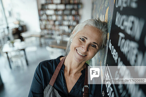 Smiling female cafe owner wearing apron leaning on blackboard in coffee shop