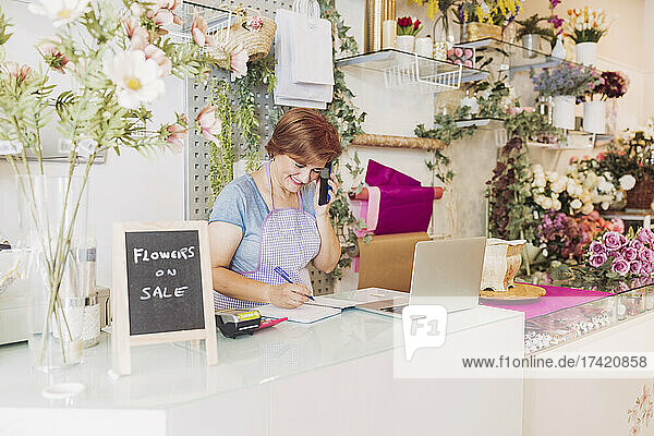 Smiling female flower shop owner talking on smart phone while writing in diary at checkout