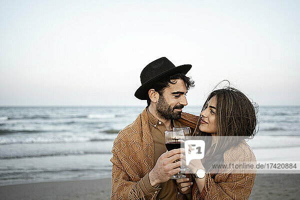 Couple wrapped in blanket toasting wineglass at beach during sunset