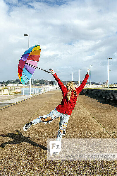 Cheerful woman holding umbrella jumping on footpath during sunny day