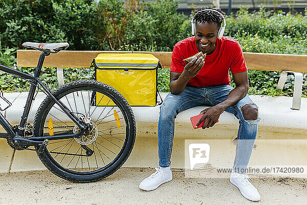 Smiling young delivery man eating food while sitting by bicycle on bench