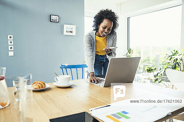 Young businesswoman working on laptop while sending voicemail through smart phone at home office
