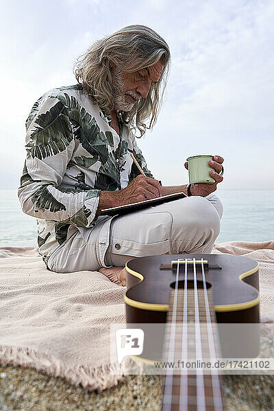 Mature man writing in book while sitting by ukulele at beach