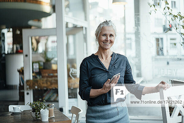 Lächelnde Freiberuflerin mit Mobiltelefon steht im Café