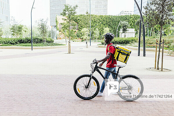 Delivery man wearing helmet while wheeling bicycle on footpath