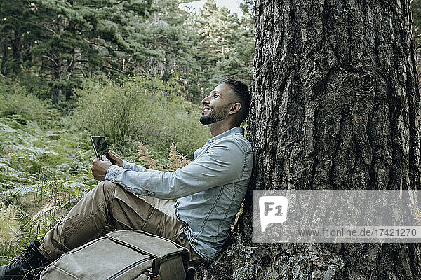 Thoughtful man holding digital tablet while leaning on tree trunk by backpack