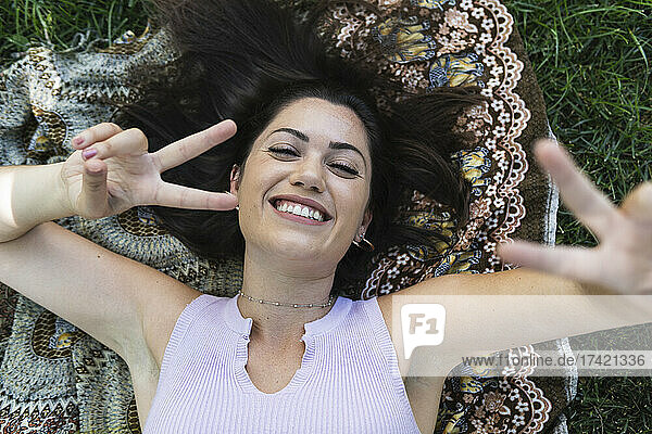 Happy young woman gesturing peace sign while lying at public park