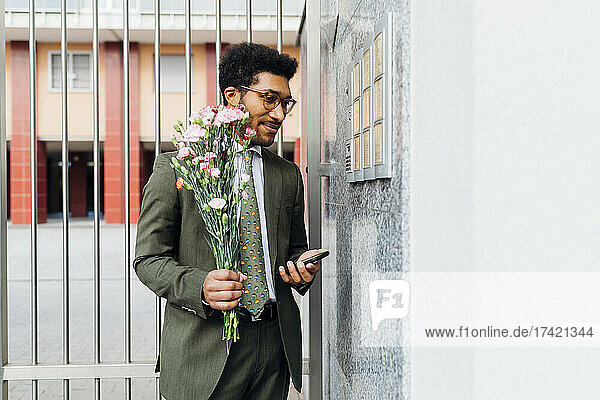 Businessman holding bouquet and mobile phone while standing by wall