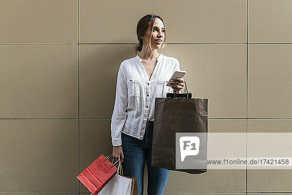 Young woman holding smart phone and paper shopping bags in front of wall