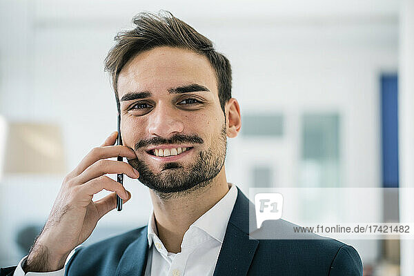 Lächelnder  gutaussehender männlicher Berufstätiger mit Haarstoppeln  der im Büro mit dem Mobiltelefon spricht