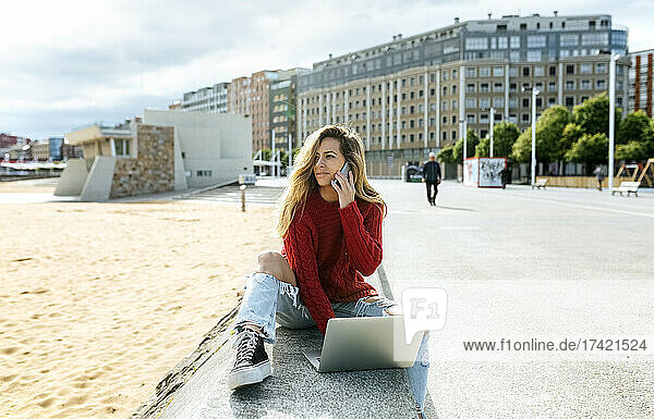 Smiling woman talking on phone while using laptop on retaining wall