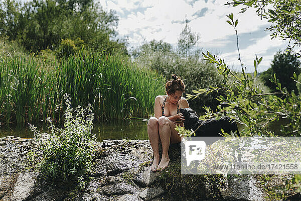 Tierbesitzerin streichelt Hund  während sie auf einem Felsen an einem Teich sitzt