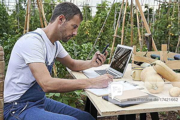 Männlicher Landarbeiter schaut auf sein Smartphone  während er am Tisch auf dem Klemmbrett schreibt
