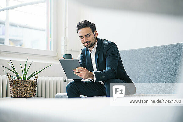Junger Geschäftsmann liest digitales Tablet und hält Kaffeetasse in der Hand  während er auf dem Sofa im Büro sitzt