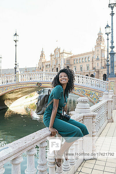 Smiling young woman with curly hair sitting on railing at Plaza De Espana  Seville  Spain