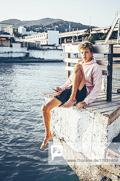 Young man holding mobile phone while sitting at pier