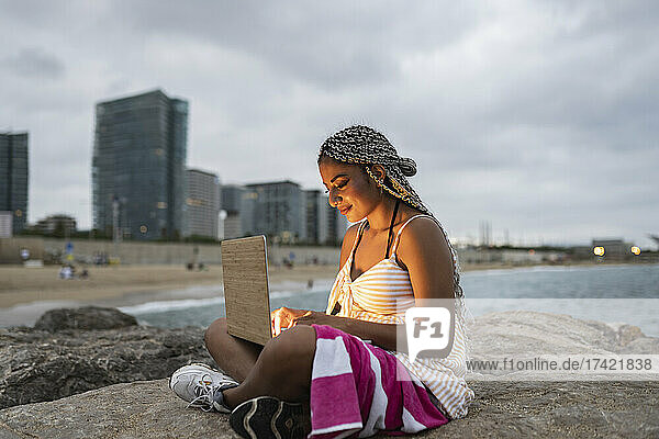 Junge Frau benutzt Laptop  während sie bei Sonnenuntergang auf einem Felsen am Strand sitzt