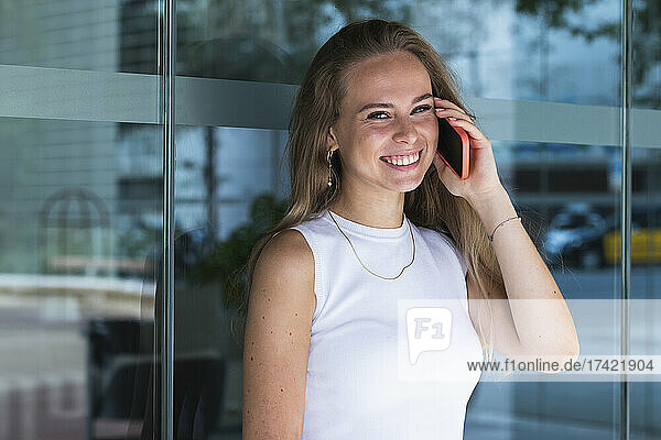 Happy young female freelancer talking on mobile phone in front of glass wall