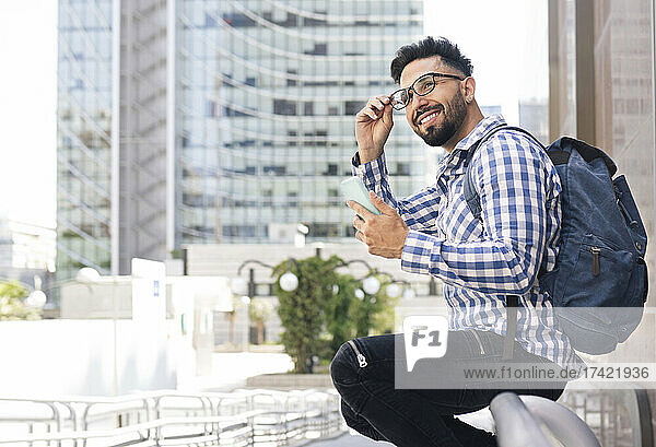 Smiling man holding eyeglasses and mobile phone sitting on railing