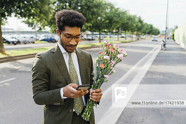 Businessman with bouquet using mobile phone while standing on street