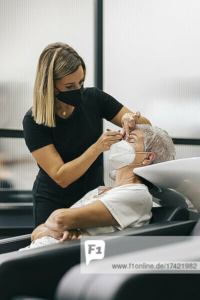 Female beautician with face mask tweezing customer's eyebrows in beauty spa