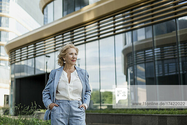 Senior businesswoman standing with hands in pockets by office building